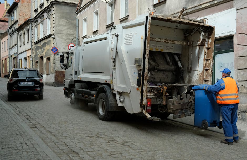A waste collection operative, dressed in a high-visibility yellow and red uniform, is positioned at the rear of a large red garbage truck, which is parked on the side of a paved street. The worker app