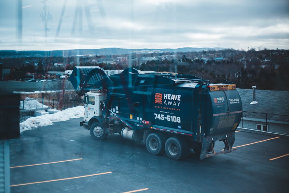 A large black waste collection truck operated by Heave Away Waste Removal is parked in an outdoor lot during overcast weather with a cloudy sky. The truck features the company's logo and contact numbe